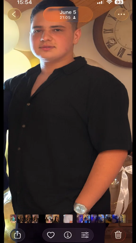 A screenshot of a photo showing a man wearing a black short-sleeved button-up shirt and a silver watch.